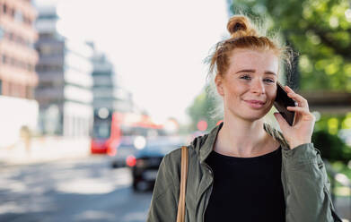 Front view portrait of cheerful young woman with red hair outdoors in town, using smartphone. - HPIF15448