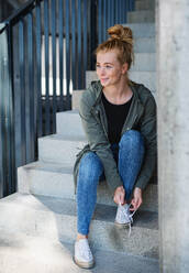 Portrait of young woman with red hair outdoors in town, sitting and tying shoelaces on staircase. - HPIF15451