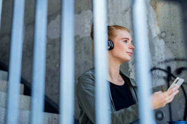 Porträt einer jungen Frau mit roten Haaren im Freien in der Stadt, die ein Smartphone und Kopfhörer benutzt. - HPIF15452