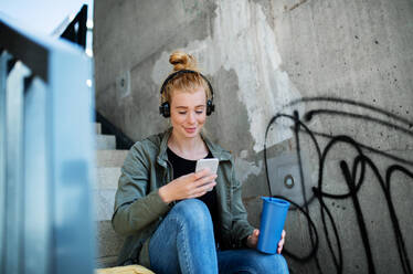 Porträt einer jungen Frau mit roten Haaren im Freien in der Stadt, die ein Smartphone und Kopfhörer benutzt. - HPIF15453