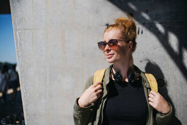 Front view portrait of cheerful young woman with red hair outdoors in town, wearing sunglasses. - HPIF15460
