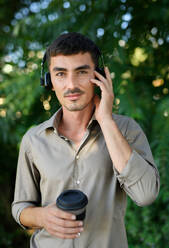 Junger attraktiver Mann mit Kopfhörern und Kaffee in einem wiederverwendbaren Becher im Freien in der Stadt. - HPIF15465