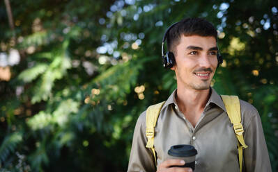 Junger attraktiver Mann mit Kopfhörern und Kaffee in einem wiederverwendbaren Becher beim Spaziergang im Freien in der Stadt. - HPIF15466