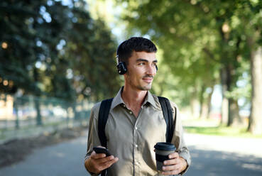 Junger attraktiver Mann mit Kopfhörern und Kaffee in einem wiederverwendbaren Becher beim Spaziergang im Freien in der Stadt. - HPIF15468