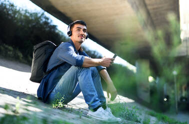 Porträt eines jungen attraktiven Mannes mit Kopfhörern und Smartphone, der in der Stadt im Freien sitzt. - HPIF15473