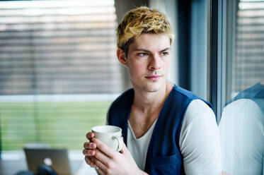 Young man with cup of coffee indoors at home, standing by window and looking out. - HPIF15598