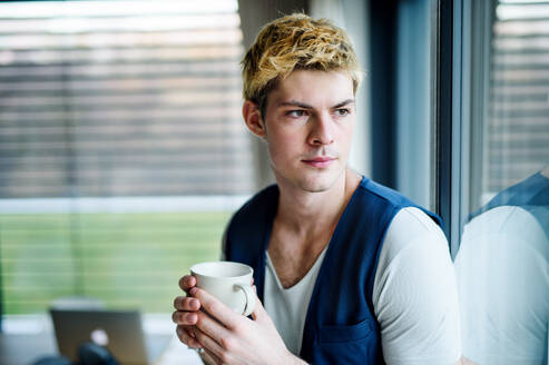 Junger Mann mit einer Tasse Kaffee zu Hause, der am Fenster steht und hinausschaut. - HPIF15598