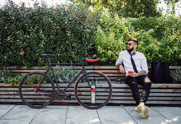 Porträt eines jungen Mannes, der mit seinem Fahrrad in der Stadt im Freien sitzt und sein Smartphone benutzt. - HPIF15601