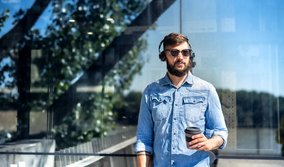 Junger attraktiver Mann mit Kopfhörern und Kaffee in einem wiederverwendbaren Becher beim Spaziergang im Freien in der Stadt. - HPIF15605