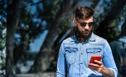 Portrait of young man with headphones standing outdoors in city, using smartphone. - HPIF15607