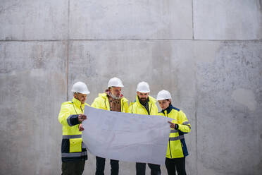 A group of engineers standing against concrete wall on construction site, holding blueprints. Copy space. - HPIF15610