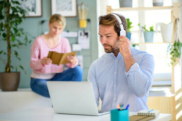 Mature businessman with headphones and laptop indoors in home office, working. Wife in the background. - HPIF15685