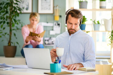 Front view of mature businessman with headphones and laptop indoors in home office, working. - HPIF15686