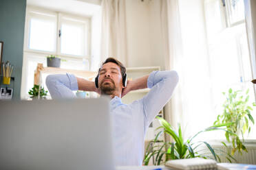 Tired mature businessman with headphones and laptop indoors in home office, resting. - HPIF15692