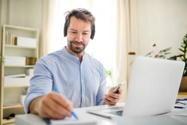 Front view of mature businessman with headphones and laptop indoors in home office, working. - HPIF15693