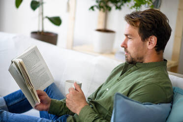 Glücklicher Mann sitzt auf dem Sofa und liest ein Buch zu Hause, Lockdown-Konzept. - HPIF15695