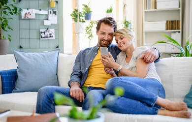 Front view of happy couple in love sitting indoors at home, using smartphone. - HPIF15713