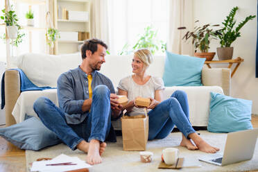 Front view of happy couple sitting on floor indoors at home, eating hamburgers. - HPIF15724