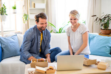 Front view of happy couple with hamburgers sitting on sofa indoors at home, using laptop. - HPIF15726