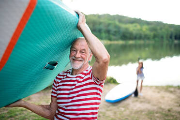 Porträt eines älteren Mannes, der im Sommer ein Paddelbrett am See trägt und in die Kamera schaut. - HPIF15763