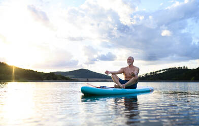 Senior man on paddleboard on lake in summer, doing yoga meditation exercise. - HPIF15788