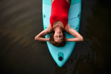 Draufsicht auf eine zufriedene ältere Frau, die im Sommer auf einem Paddelbrett auf einem See liegt und sich ausruht. - HPIF15805