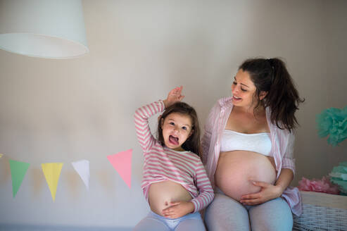 Porträt einer glücklichen schwangeren Frau mit kleiner Tochter zu Hause, die ihren Bauch zeigt. - HPIF15868