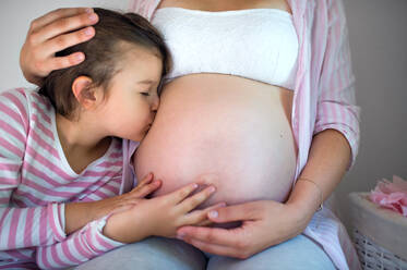Seitenansicht eines kleinen Mädchens, das den Bauch einer nicht erkennbaren schwangeren Mutter zu Hause küsst. - HPIF15870