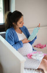 Porträt einer glücklichen schwangeren Frau zu Hause, die Babykleidung in der Hand hält und anschaut. - HPIF15884