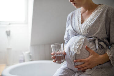 Mittelteil einer nicht erkennbaren schwangeren Frau im Badezimmer zu Hause, die ein Glas Wasser hält. - HPIF15891