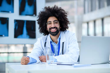 Frontalansicht eines fröhlichen männlichen Arztes, der im Krankenhaus sitzt und in die Kamera schaut. - HPIF16145