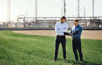 Zwei junge Ingenieure stehen im Freien bei einer Ölraffinerie und diskutieren über Probleme. - HPIF16173
