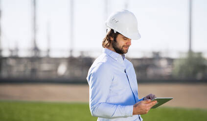 Young engineer with hard hat standing outdoors by oil refinery, using tablet. Copy space. - HPIF16176