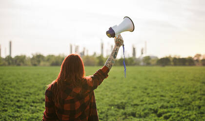 Rear view of young woman activist with megaphone standing outdoors by oil refinery, protesting. - HPIF16179