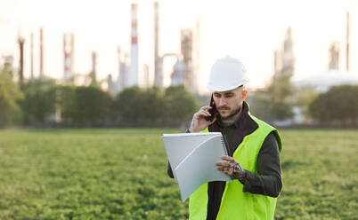 Young engineer with smartphone standing outdoors by oil refinery, making phone call. Copy space. - HPIF16187