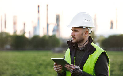 Serious young engineer with tablet standing outdoors by oil refinery. Copy space. - HPIF16188