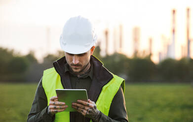 Serious young engineer with tablet standing outdoors by oil refinery. Copy space. - HPIF16189