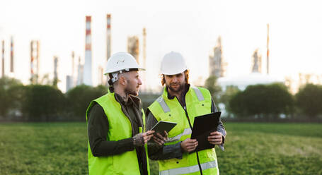 Front view of two young engineers with tablet standing outdoors by oil refinery, discussing issues. - HPIF16191