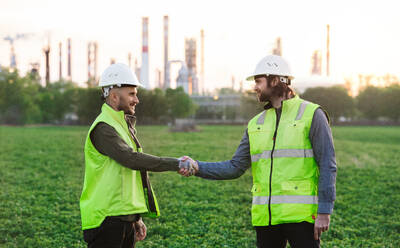 Two young engineers with hard hat standing outdoors by oil refinery, shaking hands. - HPIF16194