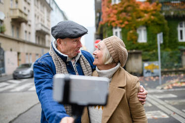 Portrait of happy senior couple walking outdoors on street in city, taking selfie. - HPIF16223
