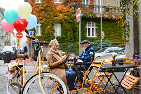 Glückliche ältere Menschen sitzen in einem Café im Freien in der Stadt und benutzen ein Smartphone. - HPIF16226