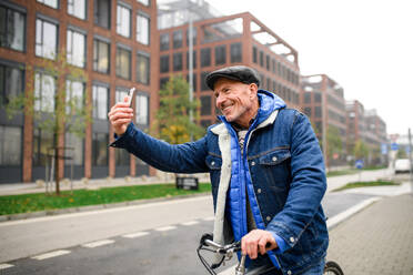 Happy senior man commuter with bicycle outdoors on street in city, taking selfie. - HPIF16246