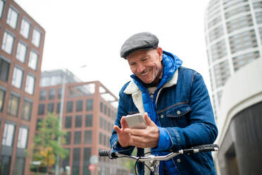 Happy senior man commuter with bicycle outdoors on street in city, using smartphone. - HPIF16247