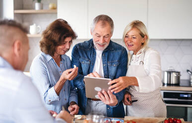 Frontansicht einer Gruppe fröhlicher älterer Freunde mit Tablet bei einer Dinnerparty zu Hause, beim Kochen. - HPIF16257