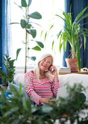 Glückliche attraktive ältere Frau mit Smartphone sitzt drinnen auf dem Sofa und telefoniert. - HPIF16299
