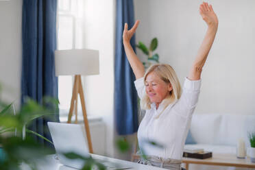 Attractive senior businesswoman with laptop sitting indoors at desk in home office, stretching when working. - HPIF16319