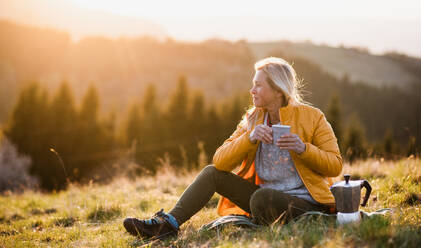 Attractive senior woman sitting outdoors in nature at sunset, relaxing with coffee. Copy space. - HPIF16359