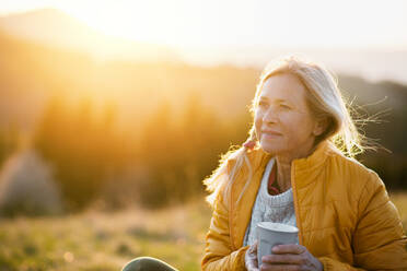 Attraktive ältere Frau sitzt im Freien in der Natur bei Sonnenuntergang, entspannt mit Kaffee. Kopieren Raum. - HPIF16360