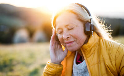 Attractive senior woman sitting outdoors in nature at sunset, relaxing with headphones. - HPIF16363