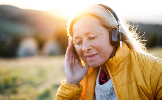Attraktive ältere Frau sitzt im Freien in der Natur bei Sonnenuntergang, entspannt mit Kopfhörern. - HPIF16363
